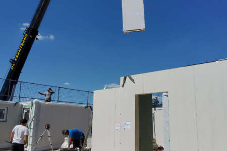 Montage einer Wand eines Fertighauses – Bauarbeiter auf Baustelle, Kran hebt Wandelement bei blauem Himmel.
