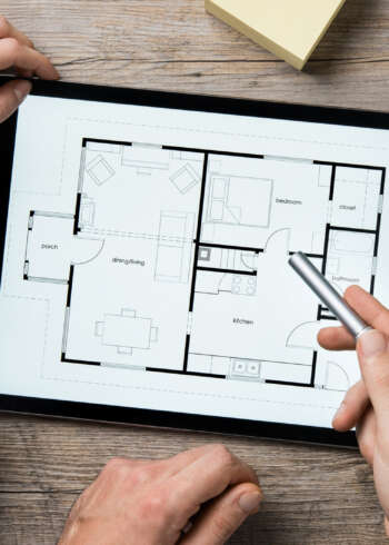 Architektinnen und Architekten besprechen Grundriss auf digitalem Tablet – Arbeitsumgebung mit Lineal, Kaffeetasse und Notizblock.