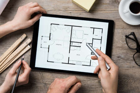 Architektinnen und Architekten besprechen Grundriss auf digitalem Tablet – Arbeitsumgebung mit Lineal, Kaffeetasse und Notizblock.