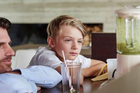 Junge sitzt mit einem Glas am Tisch und beobachtet gespannt einen Mixer mit grünem Smoothie – Moment aus dem Familienalltag in moderner Küche.