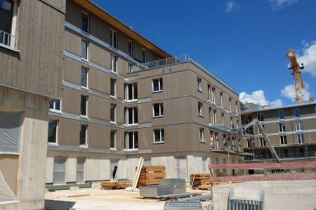 Mehrgeschossiger Hybridbau mit Holzfassade und Beton-Sockel während der Bauphase, mit Baukran im Hintergrund und Baustellenmaterial im Vordergrund.