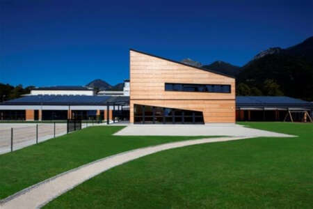 Modernes Systemgebäude mit Holzfassade und asymmetrischem Dach, gelegen vor alpiner Bergkulisse bei klarem Himmel.