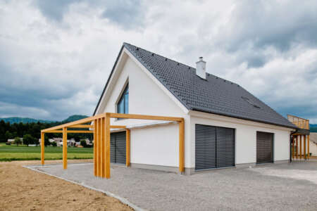 Modernes Einfamilienhaus mit Satteldach und Holzpergola, weiße Fassade und anthrazitfarbene Fensterläden.