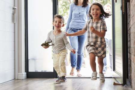 Zwei fröhliche Kinder rennen lachend durch eine geöffnete Haustür, gefolgt von einer lächelnden Frau – Momentaufnahme eines lebendigen Familienalltags beim Nachhausekommen.