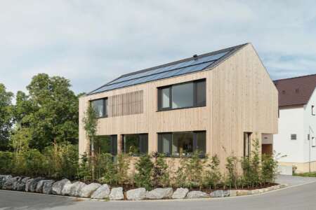 Modernes zweigeschossiges Holzhaus mit Satteldach und Photovoltaikanlage, umgeben von Natursteinen und Bepflanzung.