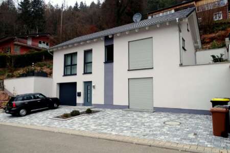 Modernes Einfamilienhaus mit Flachdach und Garage, weißer Putzfassade mit violettem Akzentstreifen, gepflasterter Einfahrt und Rollläden, gelegen in einem Hanggrundstück mit umliegenden Häusern im Hintergrund.