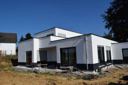 Modernes Flachdachhaus im Bau, mit weißer Fassade und schwarzen Sockelelementen, großen Glasfronten und sichtbaren Bauarbeiten im Vorgartenbereich.